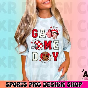 May include: A light blue t-shirt with the text "Game Day" in red, white, and black letters. The shirt features a football helmet, a football, and a hand with a peace sign.