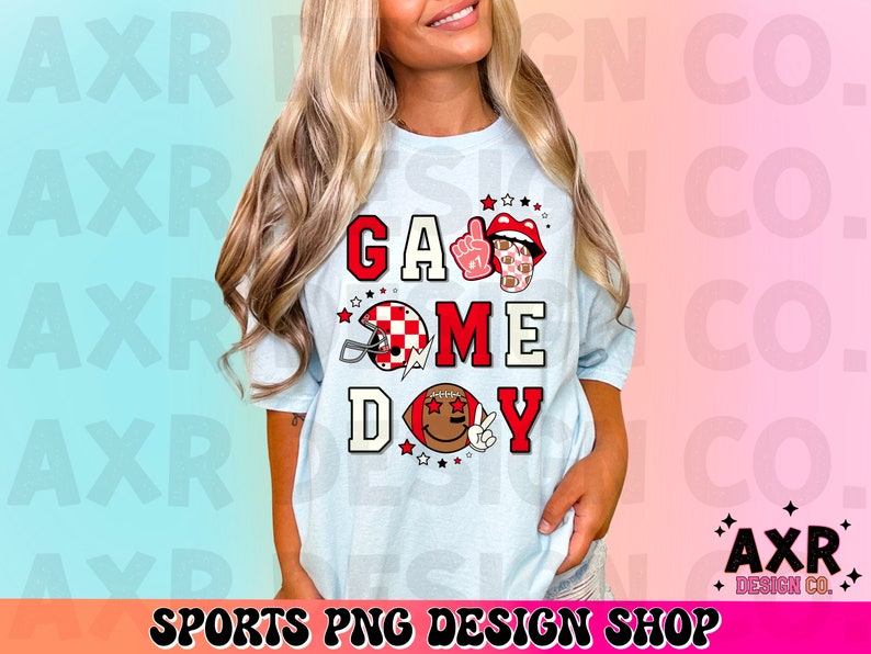 May include: A light blue t-shirt with the text "Game Day" in red, white, and black letters. The shirt features a football helmet, a football, and a hand with a peace sign.