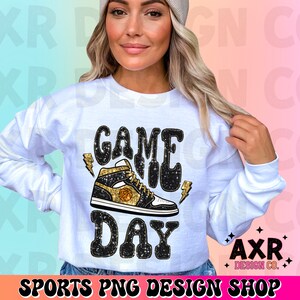 May include: White sweatshirt with the text "GAME DAY" in black glitter letters and a black and gold basketball shoe graphic. The text "AXR DESIGN CO." is in the bottom right corner.
