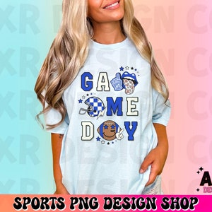 May include: A light blue t-shirt with the text "Game Day" in blue and white letters. The shirt features a football helmet, a hand giving a thumbs up, and a tongue sticking out.
