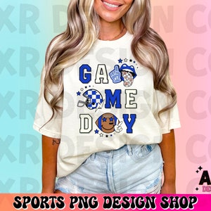 May include: White t-shirt with a blue, white, and black graphic design that says "Game Day". The graphic includes a football helmet, a football, and a hand giving a thumbs up.