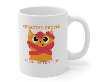 May include: White ceramic mug with a cartoon orange cat wearing a pink scarf and a pink pom-pom hat. The text "CREATIVE PEOPLE -AREN'T OFTEN TIDY- " is printed on the mug.