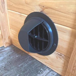 May include: A black vent with a circular trim is mounted on a light-colored wooden surface. The vent has a square-shaped outlet with a grid pattern. The wood grain is visible, and the vent is positioned near the corner of the wooden structure.