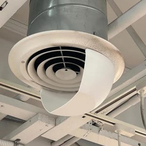 May include: A circular ceiling air diffuser featuring a metallic silver duct connection, a white outer rim, and concentric black interior rings. A curved white plastic air deflector is attached to the front, directing airflow downward.
