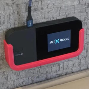 May include: A black and red MiFi X PRO 5G mobile hotspot with a screen displaying the text "MiFi X PRO 5G". The hotspot is mounted on a wall with a red plastic wall mount.
