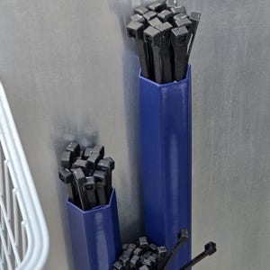 May include: Three blue, hexagonal cable tie holders with black cable ties. The holders are mounted on a silver surface. The cable ties are bundled together, ready for use.