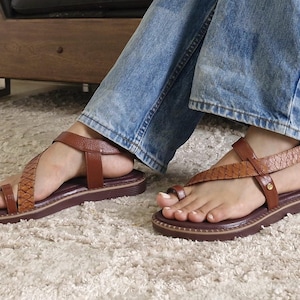 May include: Brown leather sandals with a toe ring and ankle strap. The sandals have a dark brown sole and are worn with blue jeans. The ankle strap has a woven design.