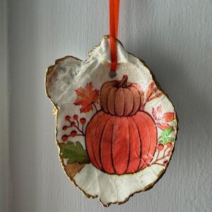 May include: A decorative ornament made from a seashell, featuring a painted design of two pumpkins and fall foliage. The shell has a gold-painted edge and is suspended by an orange ribbon.