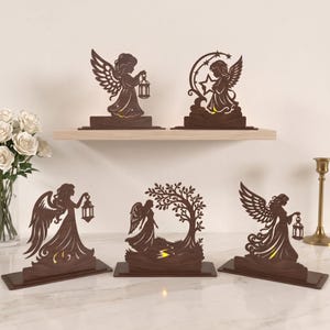 Può includere: Una collezione di cinque statuette di angeli in legno marrone scuro tagliate al laser, ciascuna montata su una base rettangolare. I disegni raffigurano angeli con ali, alcuni che reggono lanterne, altri vicino a una luna crescente, stelle o un albero, tutti illuminati da una luce calda.