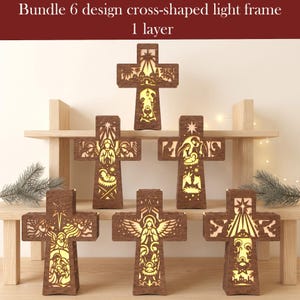 Könnte beinhalten: Ein Set aus sechs kreuzförmigen Lichtrahmen, die jeweils ein einlagiges Ausschnittdesign mit warmer Innenbeleuchtung aufweisen. Die dunkelbraunen Holzrahmen zeigen verschiedene religiöse Szenen, darunter Krippenfiguren und Engel.