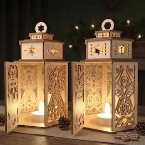 May include: Two light-coloured wooden lanterns featuring intricate laser-cut angel designs on each side. Each lantern has a square base, a tiered top with a star-shaped cutout, and an open front panel revealing a glowing white tea light candle inside.