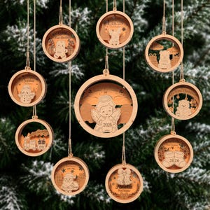 May include: A collection of round, light-coloured wooden Christmas ornaments hanging by twine. Each ornament features a layered cutout design with engraved names and holiday motifs like a gingerbread man, angel, snowman, train, and Santa.