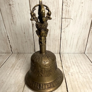 May include: A bronze-colored bell with intricate carvings. The bell features a detailed handle with a decorative top and a figure. The bell's body has ornate patterns and a flared base, showcasing a vintage aesthetic.