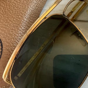 May include: Gold-tone metal sunglasses with dark green lenses and a double bridge. The inner frame is engraved with B&L 58 14 V30 IDK 5.0. The sunglasses are positioned next to a tan textured leather case with a circular logo stamp.