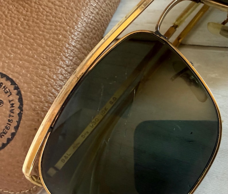May include: Gold-tone metal sunglasses with dark green lenses and a double bridge. The inner frame is engraved with B&L 58 14 V30 IDK 5.0. The sunglasses are positioned next to a tan textured leather case with a circular logo stamp.