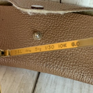 May include: Gold-tone metal sunglasses temple arm featuring the engraved text B&L 4 1/4 5 1/2 1/30 10K G.O, positioned in front of a tan textured leather carrying case with a silver metal snap button closure and a partial Ray-Ban logo visible.