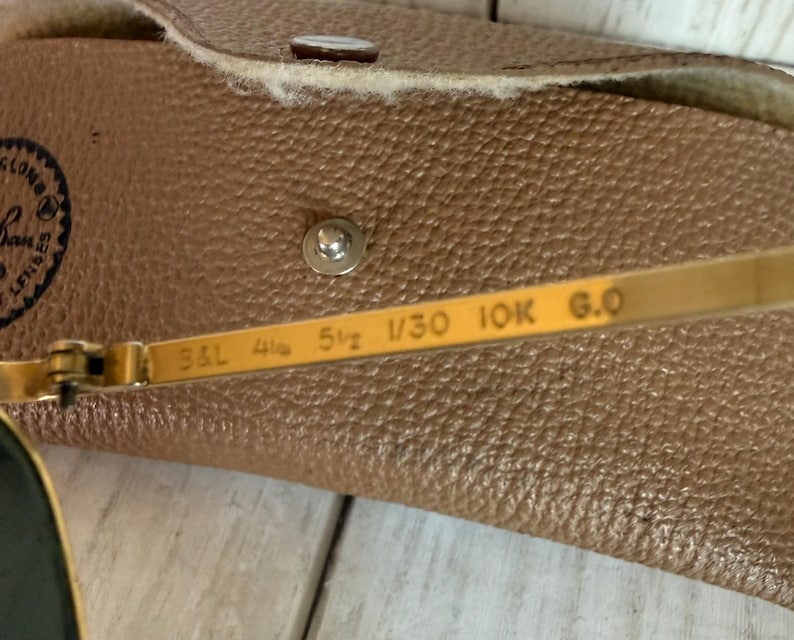 May include: Gold-tone metal sunglasses temple arm featuring the engraved text B&L 4 1/4 5 1/2 1/30 10K G.O, positioned in front of a tan textured leather carrying case with a silver metal snap button closure and a partial Ray-Ban logo visible.