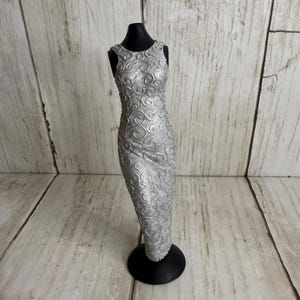 May include: A silver sleeveless dress with a swirling pattern, displayed on a black mannequin. The dress is form-fitting and reaches the ankles. The mannequin is on a black base, set against a light wood-grain background.