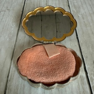 May include: An open, scalloped-edge compact case with a gold-toned metal frame and an integrated mirror. The interior contains a soft, peach-colored powder puff with a small fabric strap attached to the center for easy handling and application.