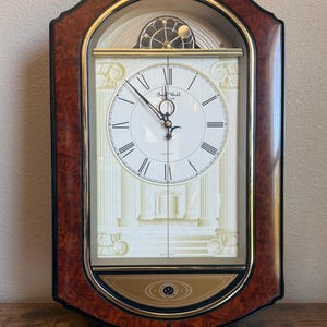 May include: An arched wall clock with a faux wood grain frame and gold trim. The white clock face features black Roman numerals, black hands, and the text Small World Rhythm. A decorative gold panel with a star motif sits at the bottom.