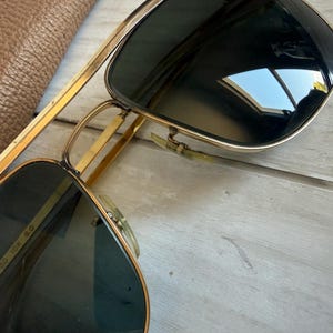 May include: Gold-tone metal aviator sunglasses featuring a double bridge design and dark tinted lenses. The inner frame arm is clearly marked with the text 10K, indicating the metal composition, along with other alphanumeric identifiers.