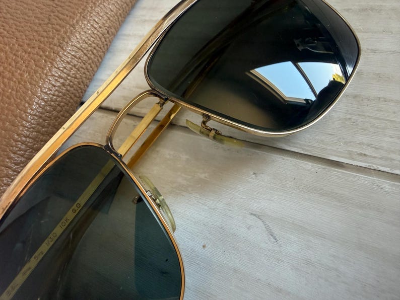 May include: Gold-tone metal aviator sunglasses featuring a double bridge design and dark tinted lenses. The inner frame arm is clearly marked with the text 10K, indicating the metal composition, along with other alphanumeric identifiers.