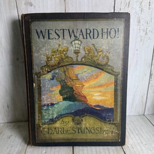 May include: Antique book titled "WESTWARD HO!" by Charles Kingsley. The cover features a colorful illustration of a sailing ship at sunset, framed by ornate gold embellishments. The book has a dark cover and shows signs of age.