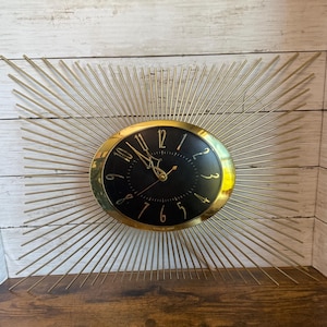 May include: A wall clock featuring a central oval black face with gold-colored numerals and hands, surrounded by a radiating sunburst design of thin gold-colored metal spokes extending outward from the center in a rectangular formation.