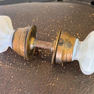 May include: Antique door knob set with white glass knobs and brass hardware. The knobs have a rounded, oval shape. The brass hardware shows signs of age and wear. The set includes two knobs, a connecting rod, and mounting plates.