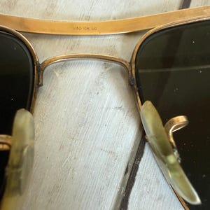 May include: Gold-tone metal aviator sunglasses featuring dark lenses and translucent nose pads. The bridge is engraved with the text 1/30 10K GO. The frame has a double bridge design with a thin upper brow bar and a curved lower bridge section.