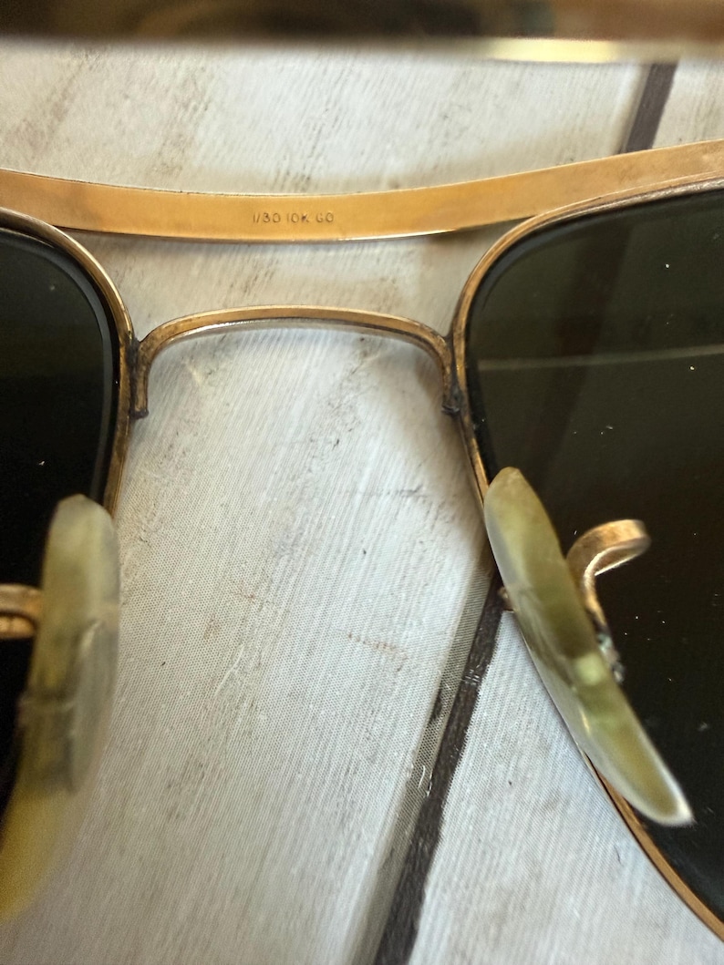 May include: Gold-tone metal aviator sunglasses featuring dark lenses and translucent nose pads. The bridge is engraved with the text 1/30 10K GO. The frame has a double bridge design with a thin upper brow bar and a curved lower bridge section.