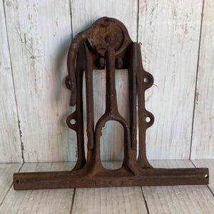 May include: A heavy, rusted cast iron mechanical component featuring a central pulley wheel, a curved lever arm, and a wide, flat base with multiple circular mounting holes. The metal surface shows significant oxidation and a dark brown texture.