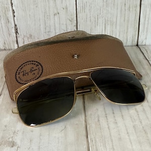 May include: Gold-tone metal sunglasses with dark lenses and a double bridge design. The sunglasses are positioned in front of a tan, textured leather-like carrying case that features a circular logo with the text Bausch & Lomb Ray-Ban.