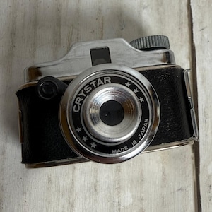 May include: A vintage camera-shaped lighter with a black leatherette body and a silver-toned metal top and lens. The lens is inscribed with "CRYSTAR" and "MADE IN JAPAN". The lighter has a textured knob and a small viewfinder.
