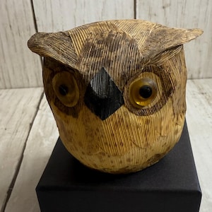 May include: A handcrafted owl figurine with a textured, layered design in shades of brown and tan. The owl has large, amber-colored eyes and a black diamond-shaped beak. The owl is sitting on a black box.