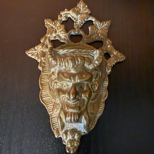 May include: A brass wall hanging featuring a devil face with horns, a goatee, and a grimacing expression. The decorative piece has a leafy crown and a cluster of grapes at the bottom. The metal has a tarnished, antique appearance.