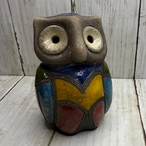 May include: A ceramic owl figurine with a patchwork design. The owl's head is gray with large, round, light-colored eyes. The body features a colorful mosaic of blue, yellow, green, and red, outlined in brown. The owl is a decorative item.