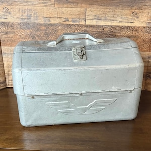 May include: A rectangular silver metal toolbox featuring a hinged lid with a central metal latch and a top handle. The front panel displays an embossed winged emblem centered horizontally. The surface shows signs of wear with minor scratches.