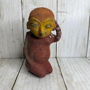 May include: A terracotta-colored figurine of a seated figure with a yellow face and closed eyes. The figure has an orange textured head and is holding its arm up to its head. The figurine has a tribal aesthetic.