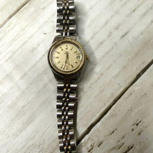 May include: A two-tone wristwatch with a gold-colored bezel and a silver and gold-tone band. The watch face is a light gold color with black hour markers and a date display at the 3 o'clock position. The watch is on a white wooden surface.