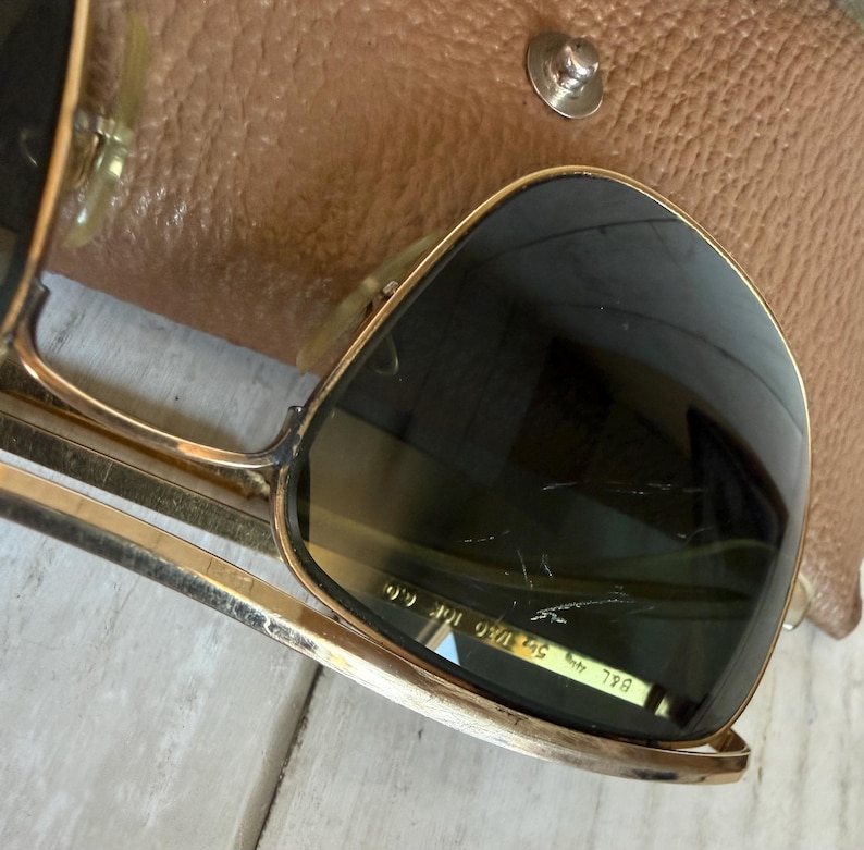 May include: Gold-tone metal sunglasses with dark lenses featuring visible surface scratches. The inner lens is etched with the text B&L 5 1/2 1/30 10K GO. The sunglasses are positioned partially over a textured tan leather case with a metal snap.