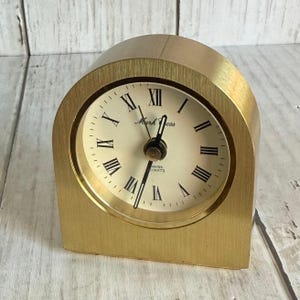 May include: A brushed gold-tone metal arch-shaped desk clock with a cream-colored face. The clock features black Roman numerals, black hour and minute hands, and the text Mark Cross and Swiss Quartz printed in black on the center of the dial.