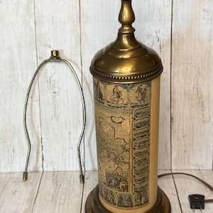 May include: Vintage brass table lamp with a cylindrical shade featuring a historical map design. The lamp has a round base and a decorative top, with a separate harp. The lamp is plugged into an electrical outlet.