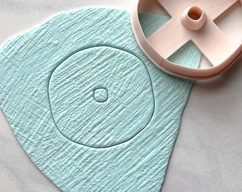 May include: A light blue, textured clay shape with a circular design and a pink cookie cutter. The cutter has an X-shaped design with a central cylinder. The clay has a rough, organic texture.