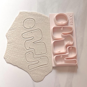 May include: A light pink plastic clay cutter with the text HMCC embossed on the side, positioned next to a piece of off-white textured clay that has been impressed with the same wavy, organic shapes as the cutter, creating a matching set.