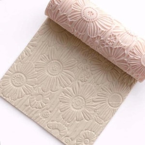 May include: A light pink rolling pin with a floral pattern, alongside a matching sheet of dough with the same embossed design. The flowers are detailed and create a textured surface, perfect for baking and crafting.