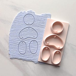 Puede incluir: Una losa de arcilla azul claro con formas recortadas para pendientes y un molde rosa con formas a juego. Las formas incluyen círculos, gotas y óvalos. Hay pequeños agujeros en las formas recortadas.