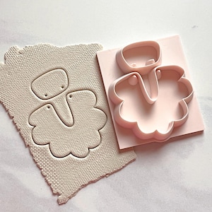 Puede incluir: Un juego de cortador y sello de pendientes de arcilla polimérica rosa claro. El cortador es una forma tridimensional del diseño del pendiente. El sello es una arcilla beige claro texturizada con el diseño del pendiente.