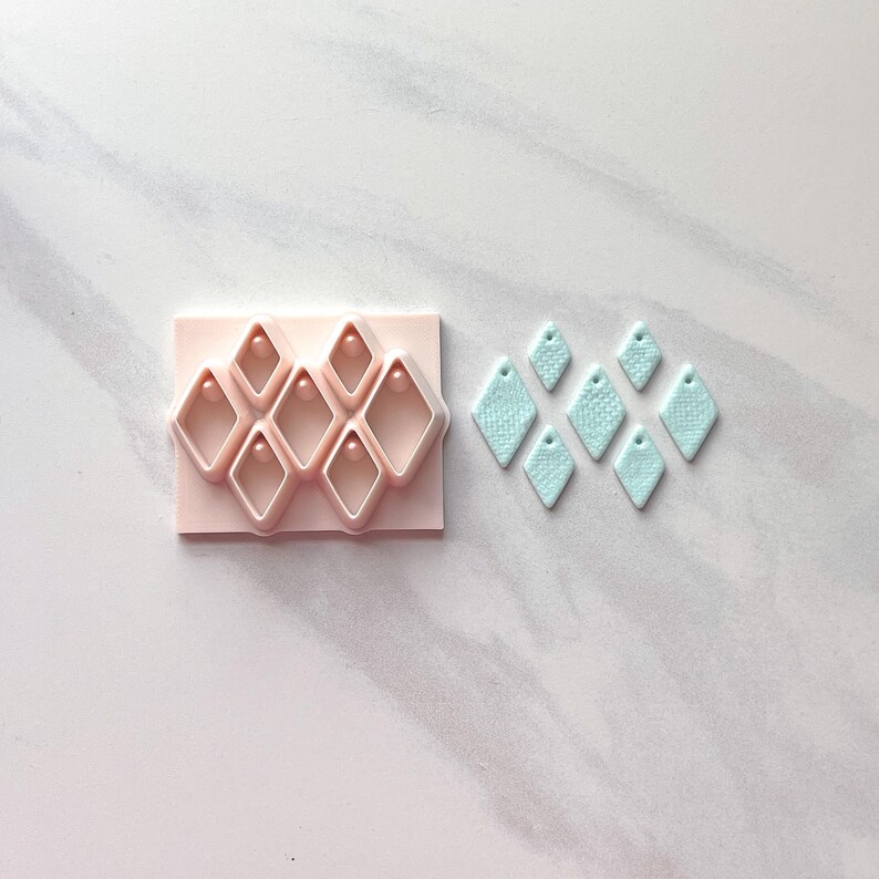 Pu&ograve; includere: Un tagliapasta rosa in argilla polimerica a forma di diamante, accanto a diversi orecchini a forma di diamante azzurro. Gli orecchini hanno una superficie strutturata e un piccolo foro nella parte superiore.
