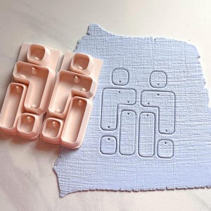 Op de afbeelding: Een lichtroze polymeerklei-uitsteker set met een familieontwerp. De set bevat twee uitstekers en een lichtblauwe, getextureerde kleilaag met de familie vorm erin gedrukt. De uitstekers hebben rechthoekige en vierkante vormen.